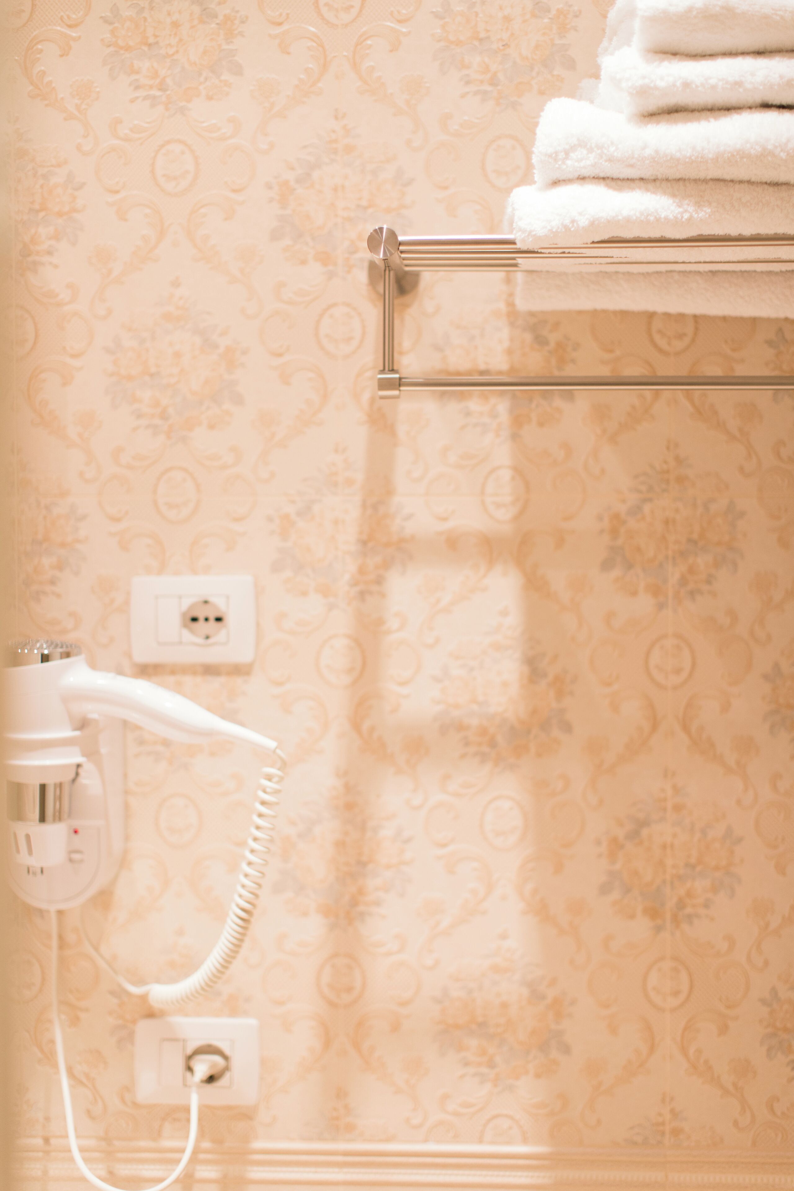 A bathroom with a patterned wallpaper, a towel rack with folded towels, and a hairdryer plugged into a wall socket.