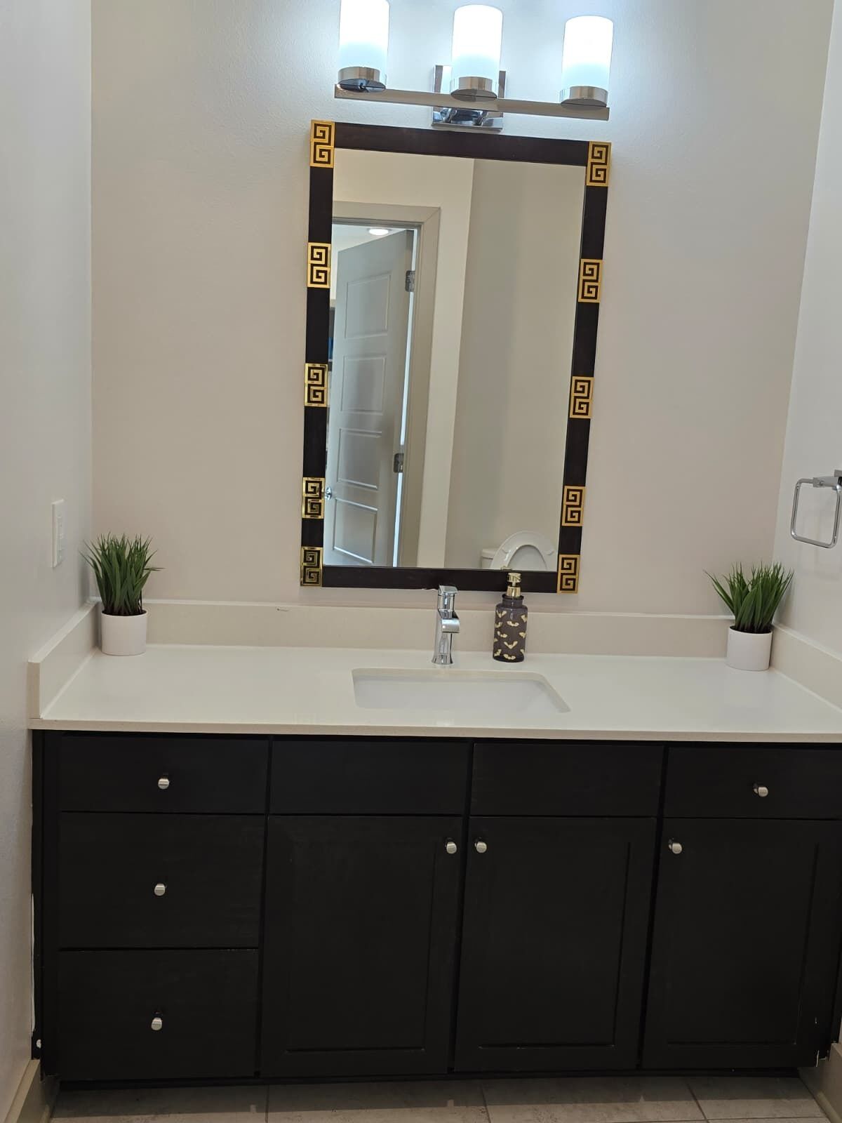 Une salle de bain avec un lavabo blanc, des meubles sombres, un miroir doté d’un cadre décoratif et deux plantes en pot.
