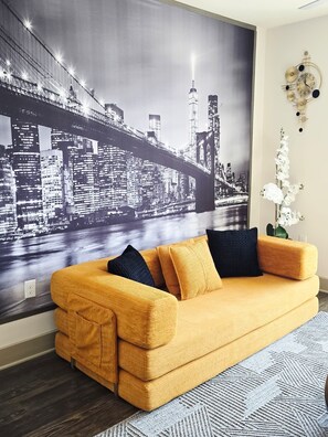 Ein Wohnzimmer mit einer großen Stadtansicht-Wandmalerei, einem senffarbenen Sofa und einer dekorativen Wanduhr.