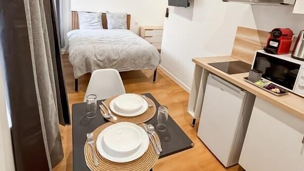 A compact hotel room with a bed, a small kitchenette, a dining area with a table set for two, and a TV mounted on the wall. - Au42BisJM (Cambrai)