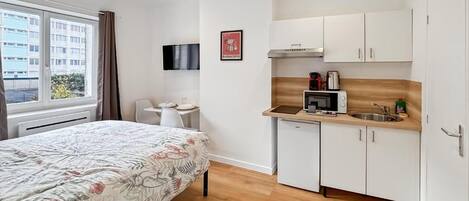 A compact hotel room with a bed, a small kitchenette, a flat-screen TV, and a window with curtains.