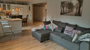 Un salon moderne avec un canapé gris, un coin repas comprenant une table et des chaises, et une cuisine équipée de meubles et d’un luminaire suspendu.