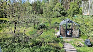 Une terrasse en bois mène à une serre en verre, elle-même entourée d’une végétation luxuriante et d’arbres.