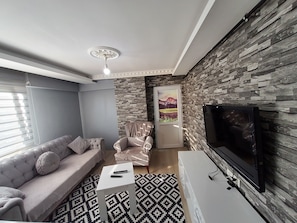 Una sala de estar moderna con un sofá gris, un tapete estampado, una mesa de centro blanca, un sillón blanco, una pared de piedra, una puerta con una pintura de paisaje y un televisor de pantalla plana.