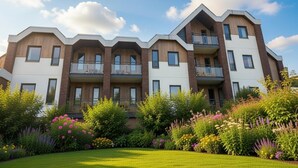 Un edificio residencial de varios pisos con balcones, un césped cuidado y una variedad de plantas con flores.
