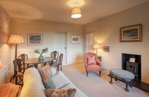 Ein gemütliches Wohnzimmer mit einer Couch, einem Sessel, einem Hocker, einem Esstisch mit Stühlen, einem Kamin und gerahmten Bildern an den Wänden.