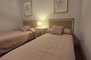Dos camas individuales con cabeceras, una mesita de noche con lámpara, y dos citas enmarcadas en la pared.