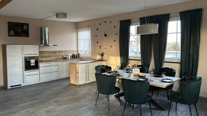 Una cocina moderna con una isla central, un comedor con mesa de madera y sillas verdes, y vistas al exterior a través de amplios ventanales.