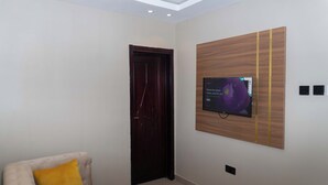 Egy modern szoba, sötét színű, fából készült ajtóval, a falra szerelt, lapos képernyős televízióval, és világos színű, sárga párnával ellátott karosszékkel.