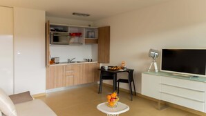 A modern living room with a sofa, a small round table with fruit, a TV, and a kitchen area with cabinets and a microwave. - Charming T1 in residence with swimming pool (Porto-Vecchio)