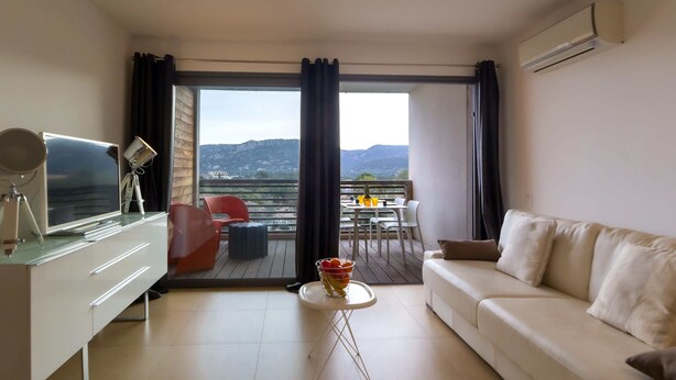 A modern living room with a white sofa, a glass coffee table, and a TV on a white cabinet. A balcony with a table and chairs is visible through the open curtains. - Charming T1 in residence with swimming pool (Porto-Vecchio)