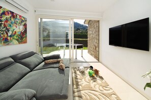 A living room with a grey sofa, a flat-screen TV, a colorful abstract painting, and a glass door leading to an outdoor area. - Villa CYN-pool, billiards, piano and miniature golf (Santo Stefano di Camastra)