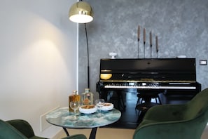 Huoneessa on piano, pyöreä pöytä, jonka päällä on lasilevy, sekä kaksi vihreää tuolia.