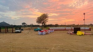 An outdoor event area with a dirt ground, a few vehicles, and a stage with a backdrop. - Shree Karni Hotel & Restaurant (Bikaner)