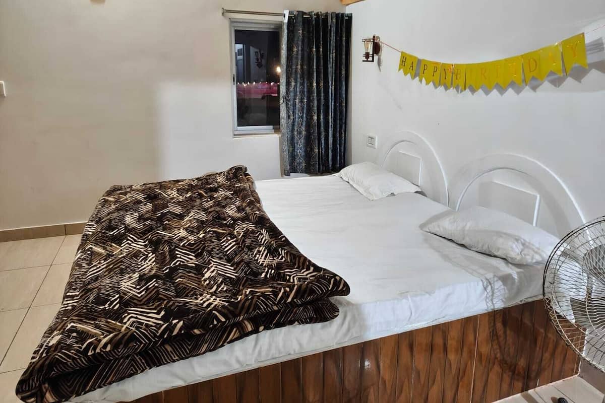 Une chambre avec un lit, une fenêtre avec des rideaux, un ventilateur et une banderole jaune sur laquelle on peut lire « JOYEUX ANNIVERSAIRE ».