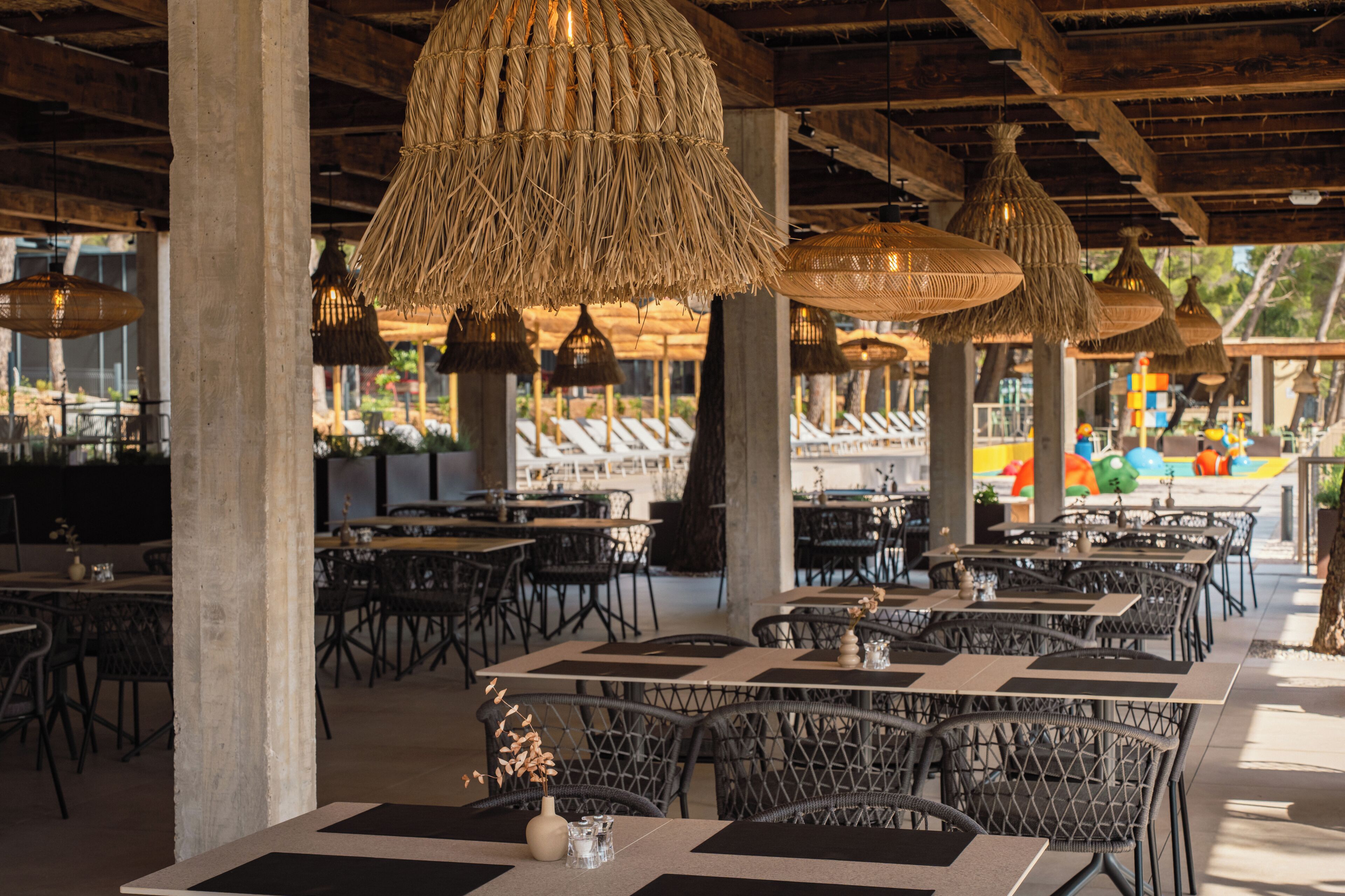An outdoor dining area with wicker tables and chairs, hanging wicker lamps, and a view of the beach in the background.