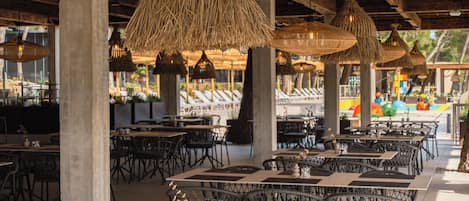 An outdoor dining area with wicker tables and chairs, hanging wicker lamps, and a view of the beach in the background.