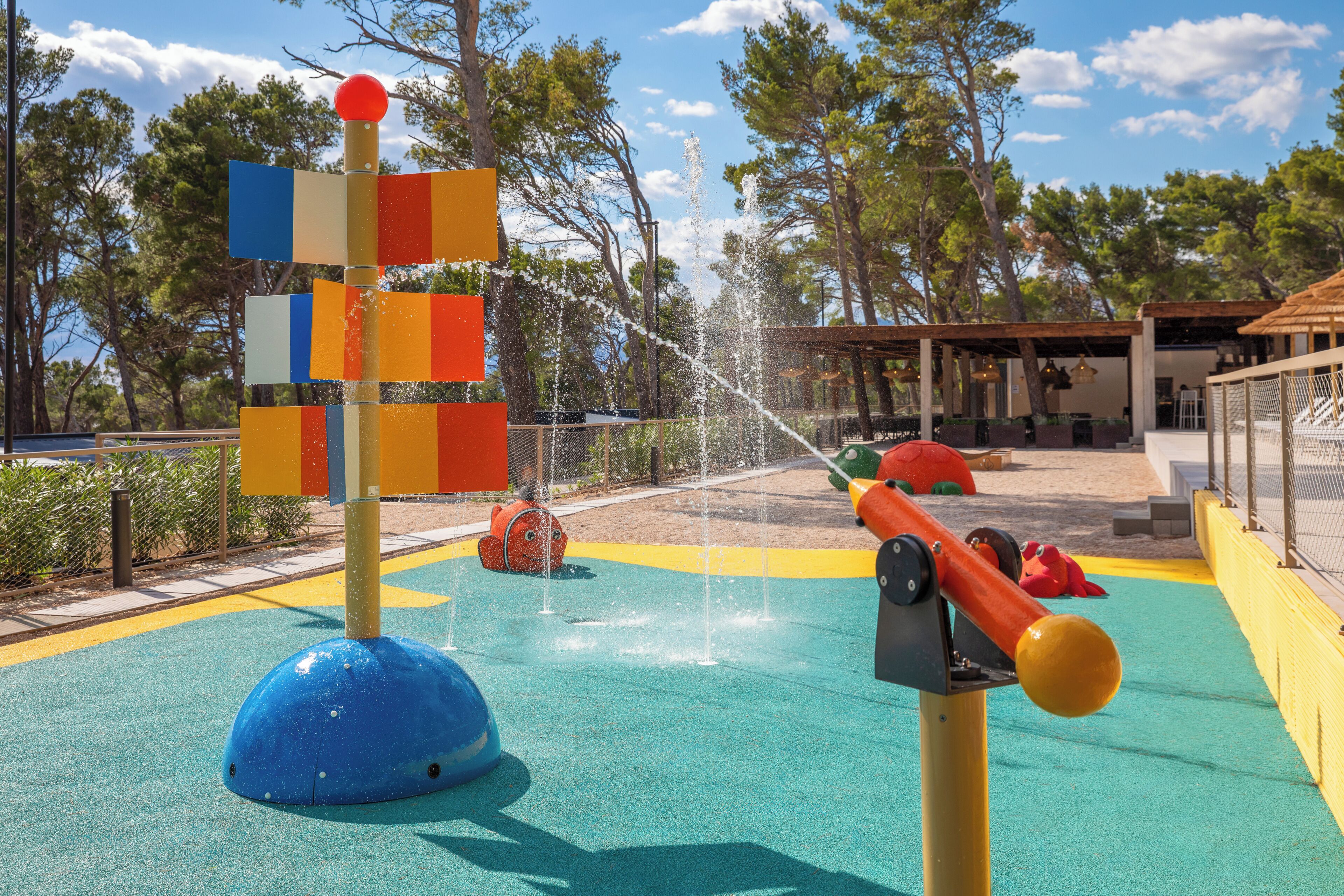 Ein Wasserspielbereich mit einer bunten Spielkonstruktion, einer Wassersäule und einem überdachten Sitzbereich.