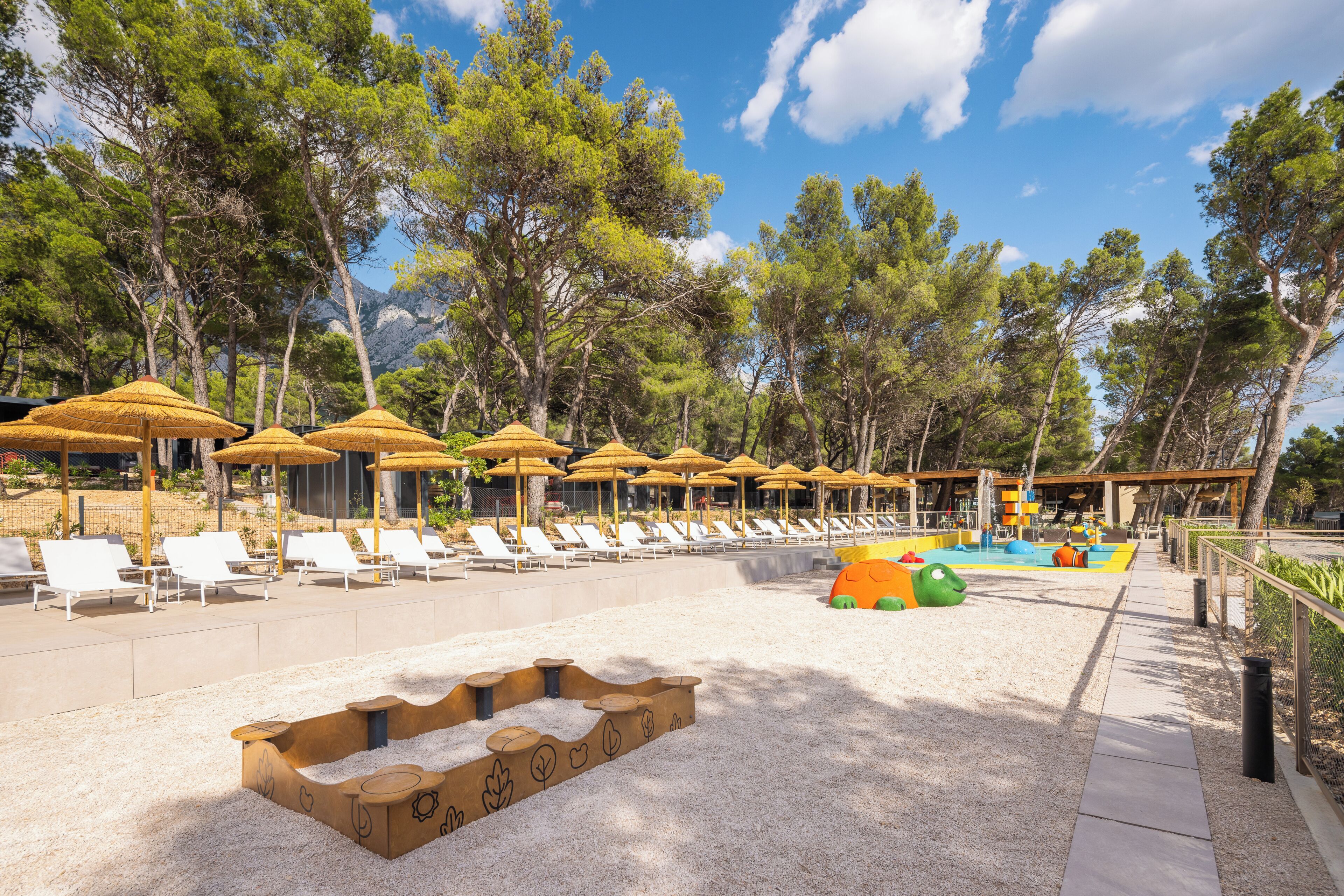 Un complexe hôtelier situé en bord de mer, équipé de chaises longues, de parasols et d’un espace de jeux pour enfants.