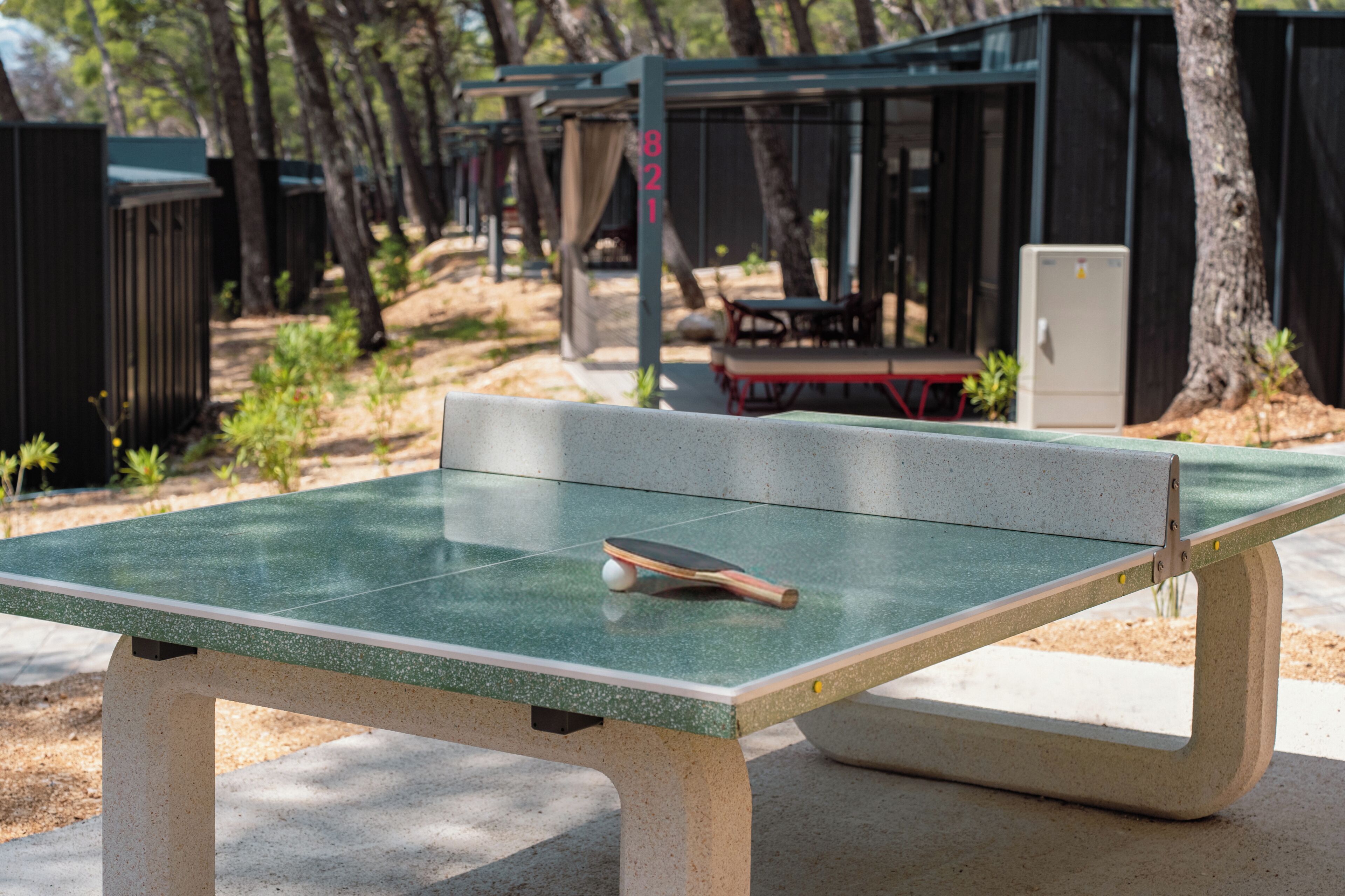 Una mesa de ping pong con una paleta y una pelota, rodeada de árboles y un área de descanso techada.
