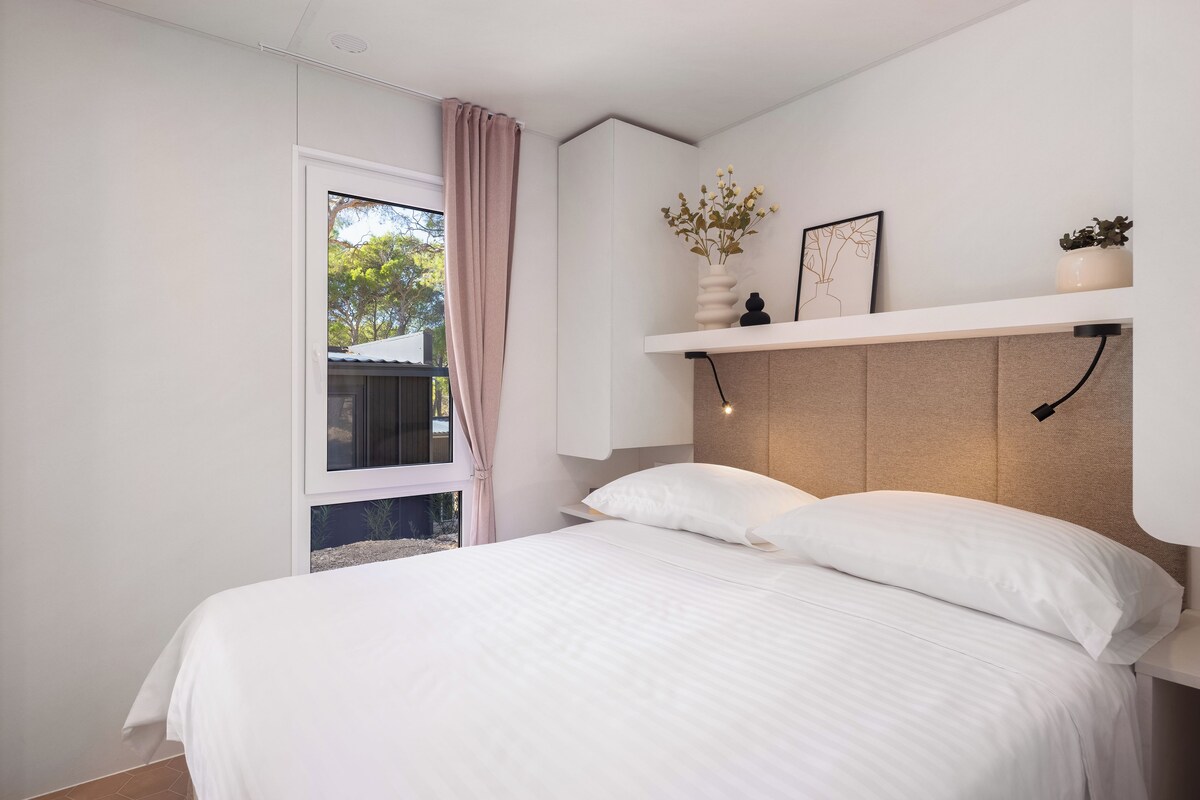A bedroom with a bed, a window with a curtain, a wall-mounted shelf with decorative items, and a headboard with a light fixture.