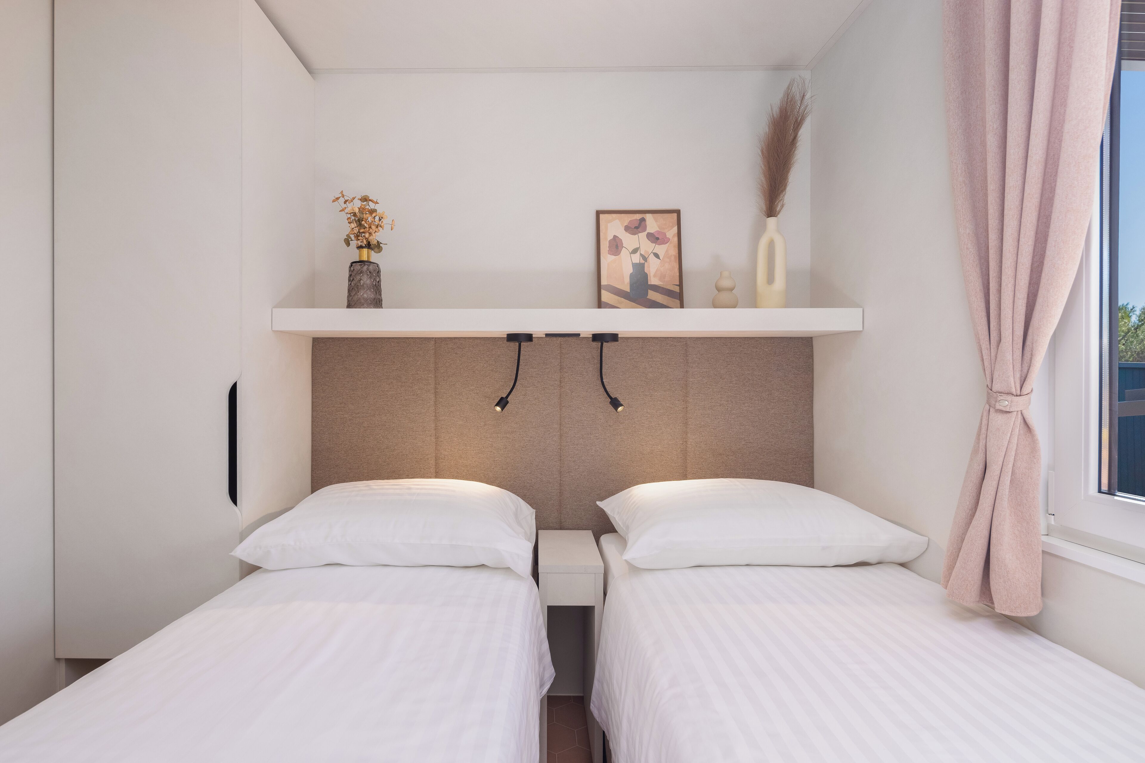 Une chambre avec deux lits simples, une étagère ornée d’objets décoratifs, une table de chevet et une fenêtre avec un rideau.