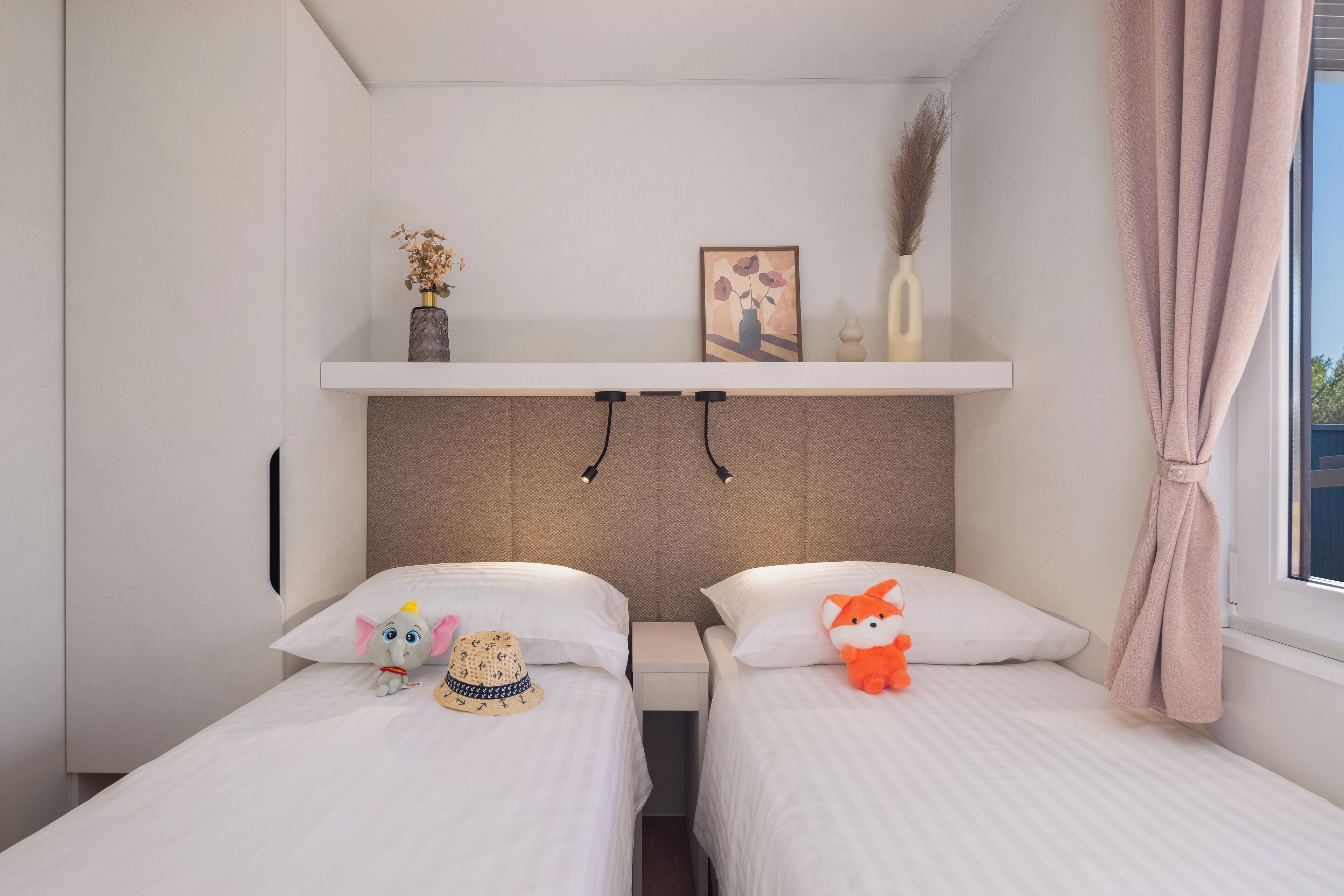 Una habitación de hotel con dos camas, un estante con adornos y una ventana con cortina.