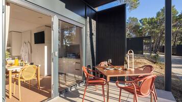 Un área de comedor al aire libre y moderna, con una mesa, sillas, un ventanal de vidrio que da a una cocina y vistas del paisaje circundante.