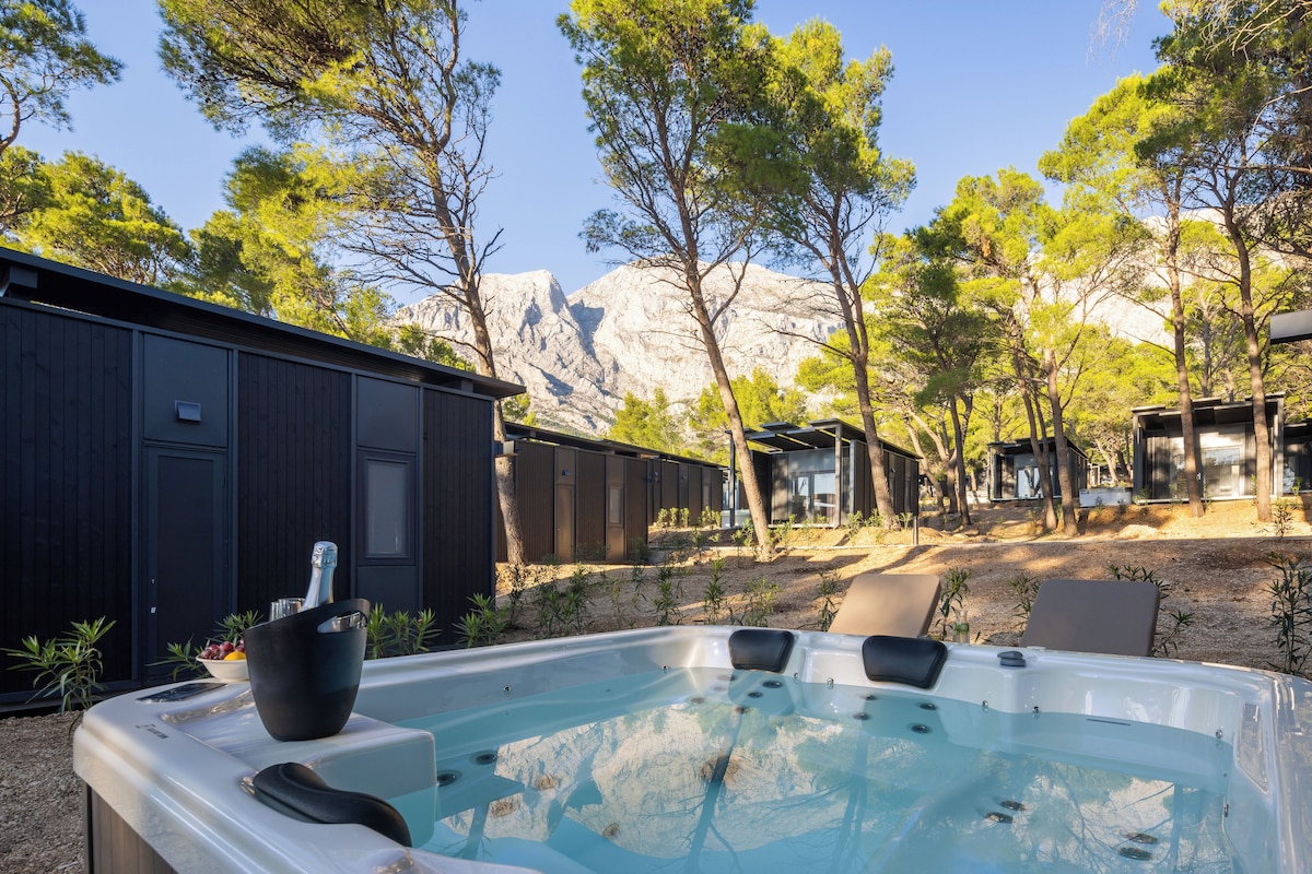 A hot tub with a mountain backdrop, surrounded by trees and outdoor seating.