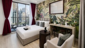 Ein Hotelzimmer mit einem großen Bett, einem Schreibtisch mit zwei Hocker, Blick auf die Stadt durch große Fenster und einem Wandgemälde im tropischen Stil.