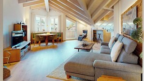 Ein großzügiger Wohnbereich mit hohen Decken und freiliegenden Balken, ein bequemes Sofa, ein Esstisch mit Stühlen und eine Küche im Hintergrund.