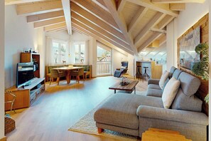 Ein großzügiger Wohnbereich mit hohen Decken und freiliegenden Balken, ein bequemes Sofa, ein Esstisch mit Stühlen und eine Küche im Hintergrund.