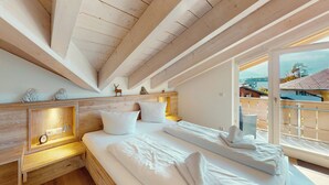 Ein Schlafzimmer mit Holzbalkendecke, einem Bett mit weißen Leinentüchern, einem Nachttisch mit einer Uhr und einem Fenster mit Blick auf Gebäude.