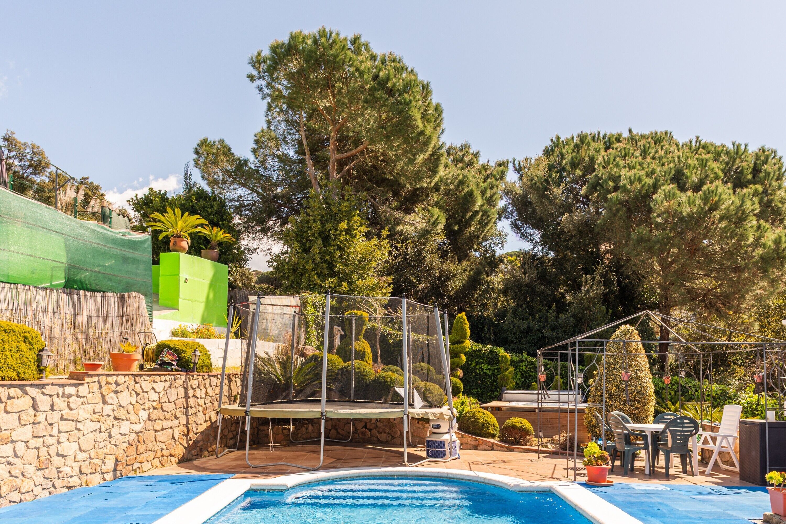 A swimming pool with a trampoline, surrounded by potted plants and a covered play area.