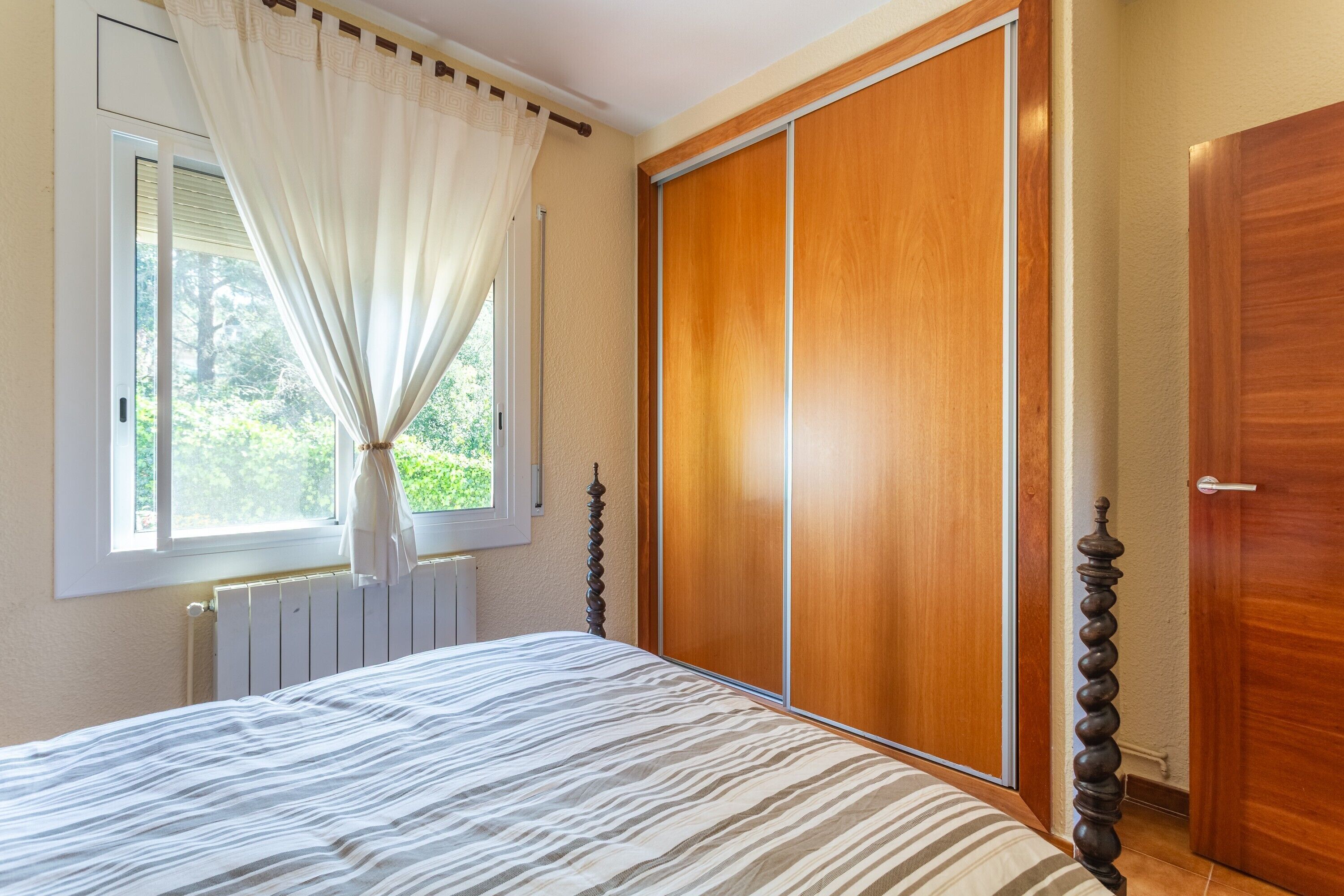 A bedroom with a bed, a window with curtains, a radiator, a wooden wardrobe, and a door.