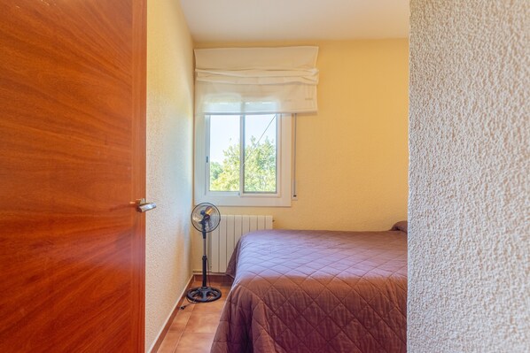 A bedroom with a wooden door, a window with a white valance, a standing fan, and a bed with a quilted cover. - Holiday Home 'Alicia y Rudi' with Wi-Fi (Vidreres)