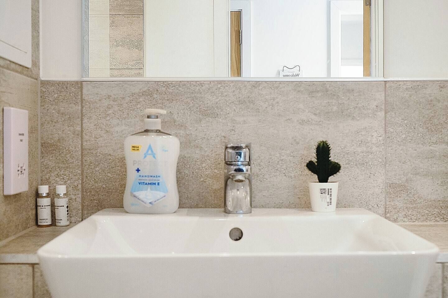 Une salle de bain avec un lavabo blanc, un flacon de lotion à la vitamine E, un salier en verre et un petit cactus en pot.