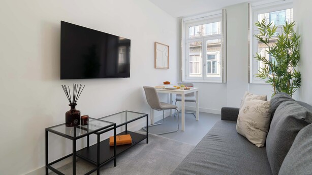 Een moderne woonkamer met een flatscreen televisie aan de muur, een glazen salontafel met een boek, een grijze bank, een eettafel met stoelen en een groot raam met uitzicht op gebouwen.