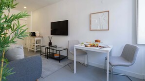 Een moderne woonkamer met een witte bank, een grijze fauteuil, een eettafel met een schaal fruit, en een aan de muur bevestigde televisie.