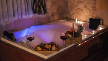 A spa tub with a wooden edge, a fruit basket, and two glasses of red wine on a marble countertop.