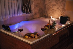 Un bain à remous bordé de bois, un panier de fruits et deux verres de vin rouge posés sur un comptoir en marbre.