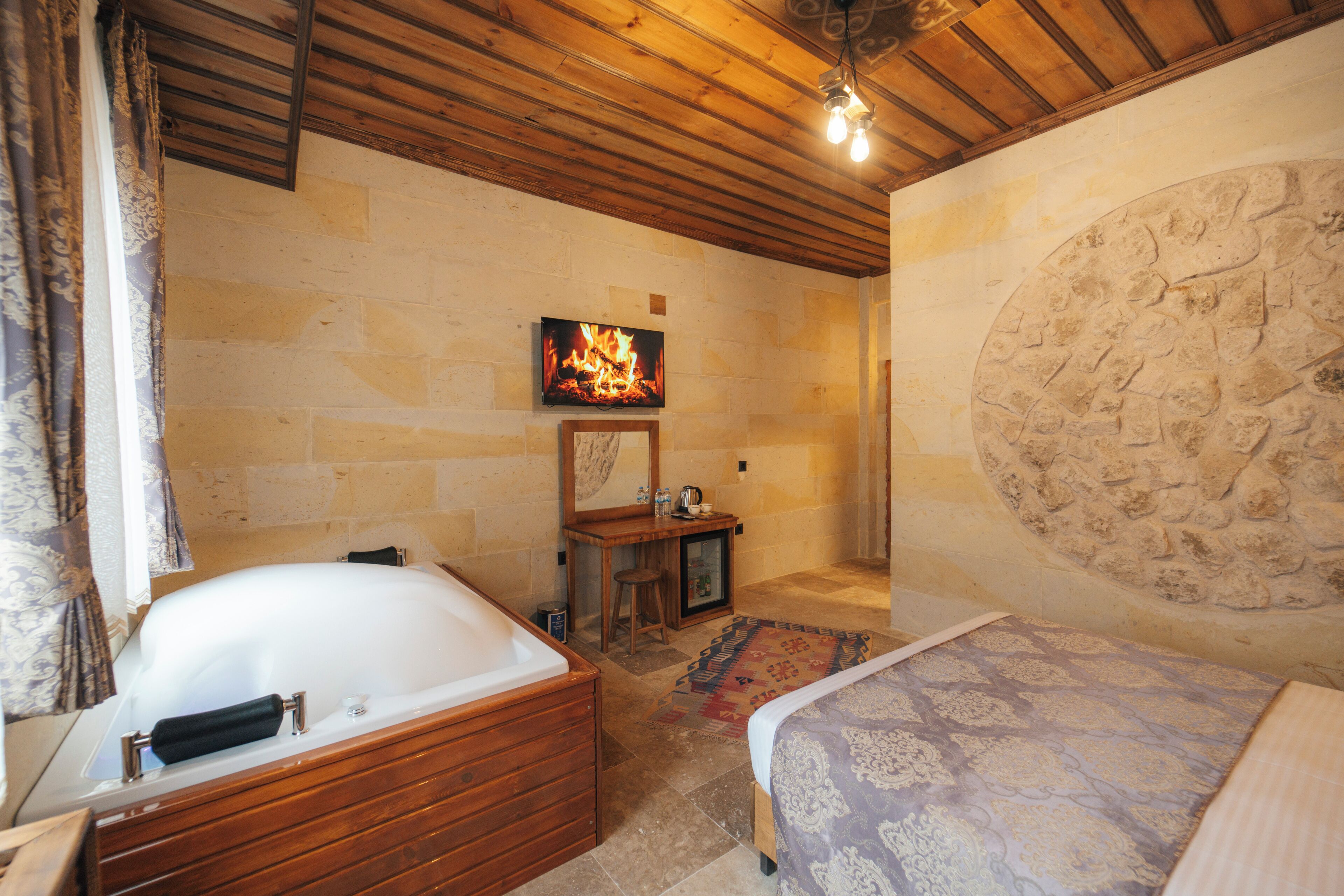 A room with a stone wall, a wooden ceiling, a bathtub, a bed, a small table with a stool, and a television displaying a fire.
