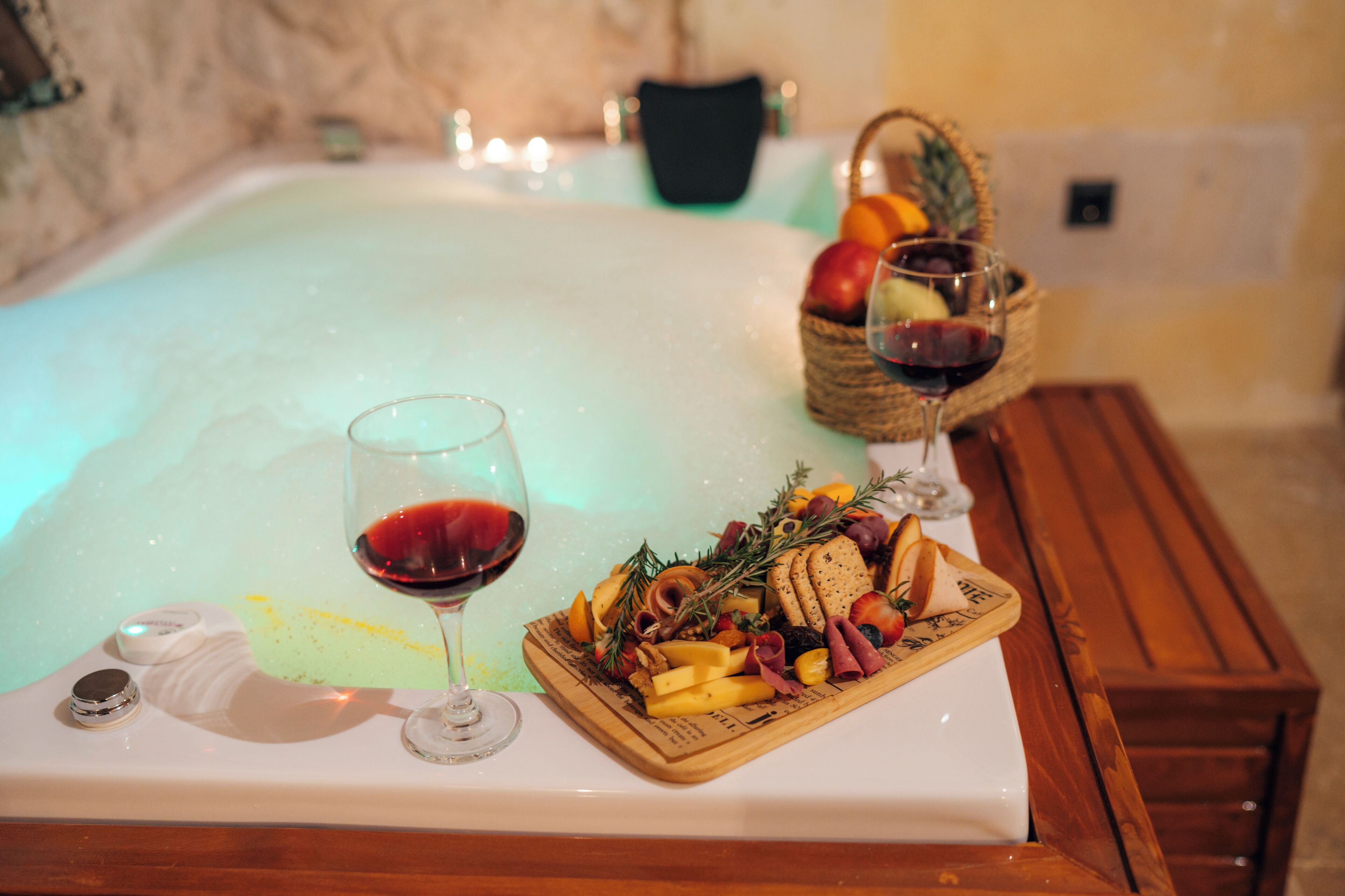 Una bañera de hidromasaje con un banco de madera, una bandeja de aperitivos y dos copas de vino tinto.