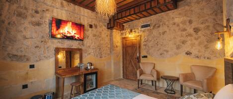 A cozy room with a bed, a desk, two chairs, a fireplace, and a stone wall.