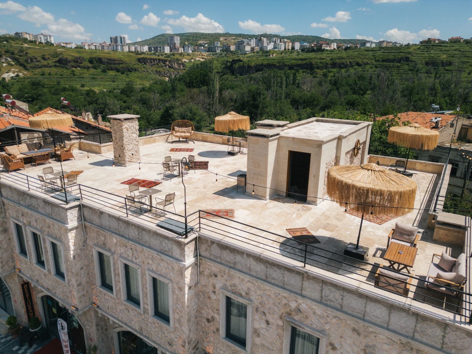 A rooftop terrace with outdoor seating, a stone building, and a view of a cityscape and hills in the background.