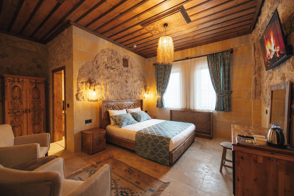 A rustic bedroom with a stone wall, wooden ceiling beams, a bed with patterned bedding, a wooden nightstand, a chair, and a dresser with a kettle.