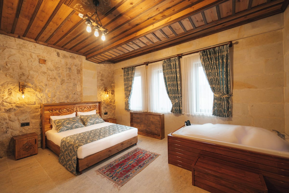 A bedroom with a stone wall, wooden ceiling, a large bed with patterned bedding, a wooden nightstand, a bedside table, a window with curtains, and a wooden bathtub.