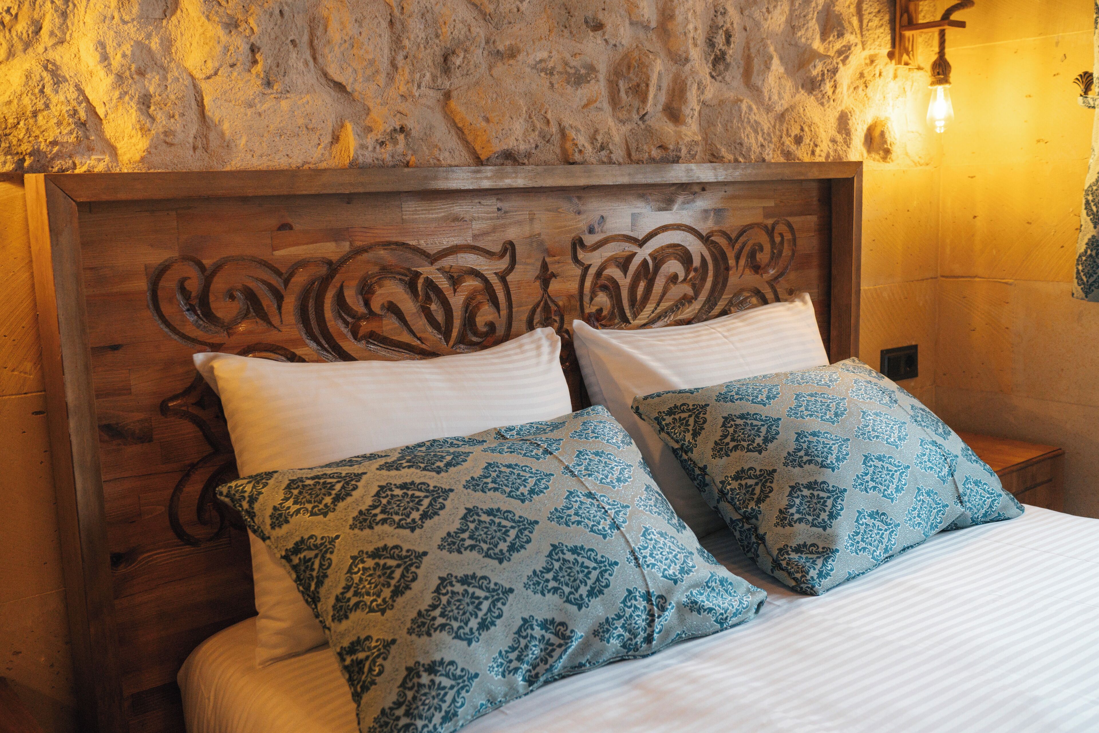 A bed with a wooden headboard, two patterned pillows, and a wall with a textured stone surface and a wall-mounted light fixture.