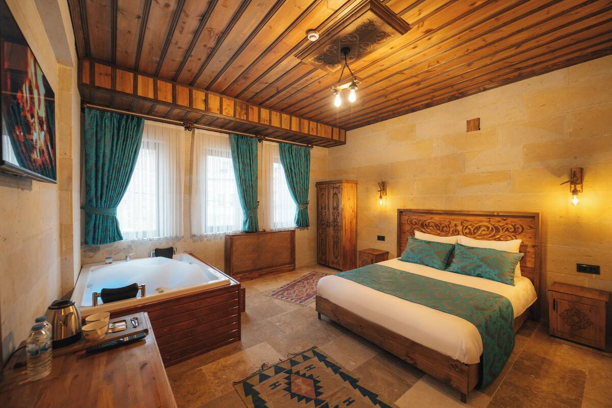 A bedroom with a wooden ceiling, a large bed with a patterned bedspread, a bathtub, and a wooden nightstand.