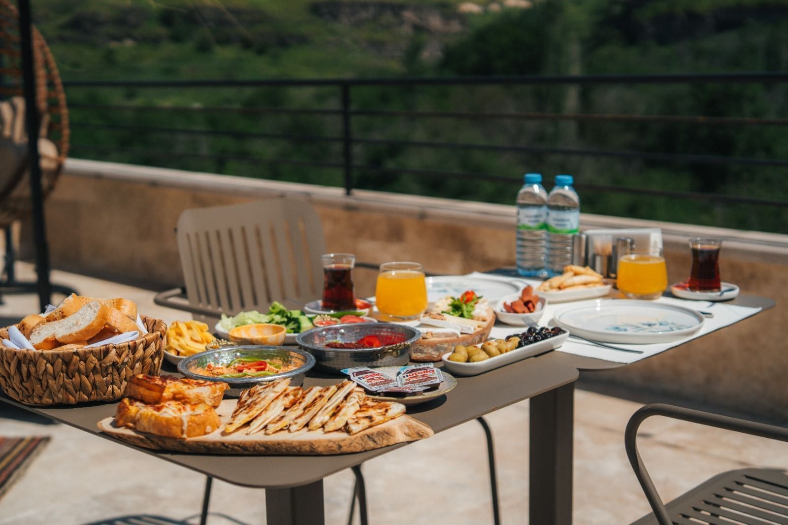 A table set with various foods and drinks, including bread, salads, and beverages, on an outdoor patio with a view of greenery.